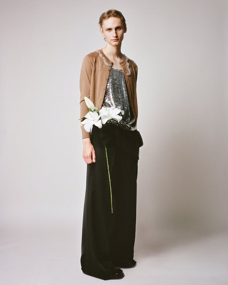 Full-length portrait of a slender person with a neutral expression, wearing a beige cardigan, shiny top, black pants, and holding white flowers.