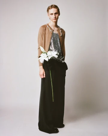 Full-length portrait of a slender person with a neutral expression, wearing a beige cardigan, shiny top, black pants, and holding white flowers.