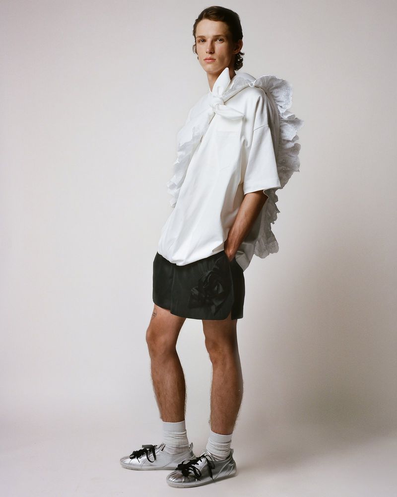 A young adult male fashion model poses indoors wearing a white, oversized shirt with ruffles, dark shorts, socks, and silver sneakers.