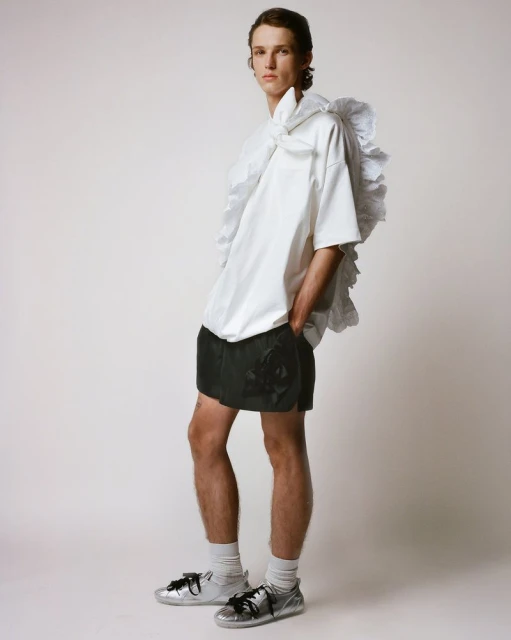 A young adult male fashion model poses indoors wearing a white, oversized shirt with ruffles, dark shorts, socks, and silver sneakers.