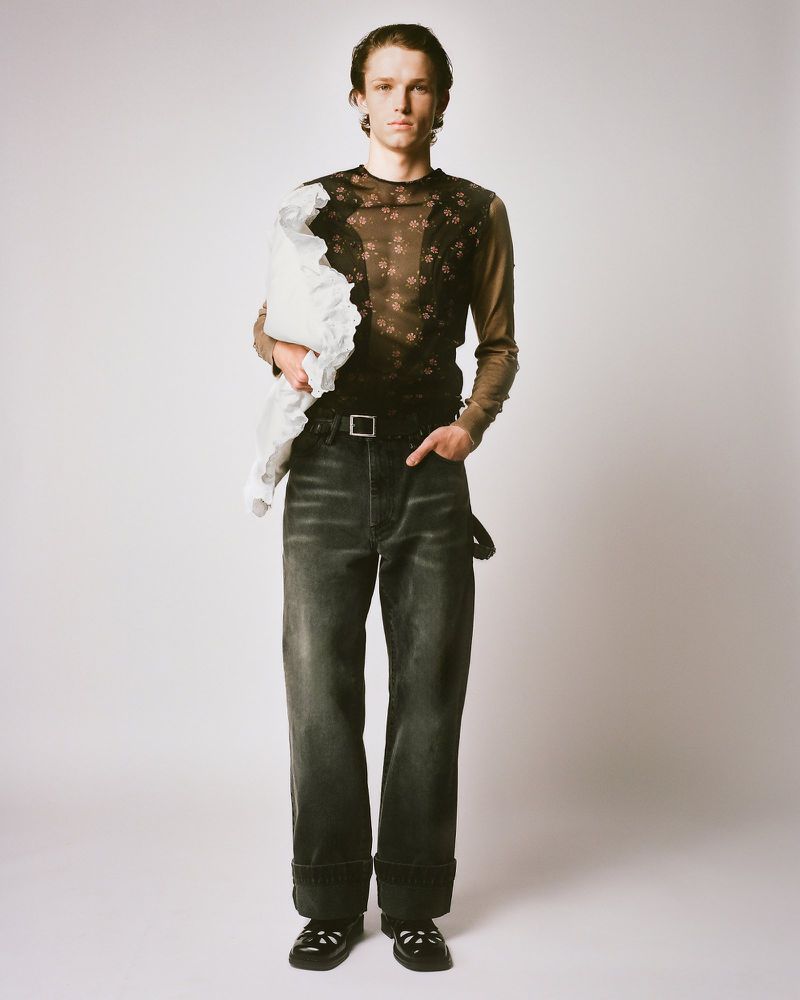 Full-length studio portrait of a young adult man in floral long-sleeve top and dark jeans, holding a white fabric, standing on a light background.