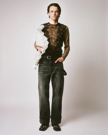 Full-length studio portrait of a young adult man in floral long-sleeve top and dark jeans, holding a white fabric, standing on a light background.
