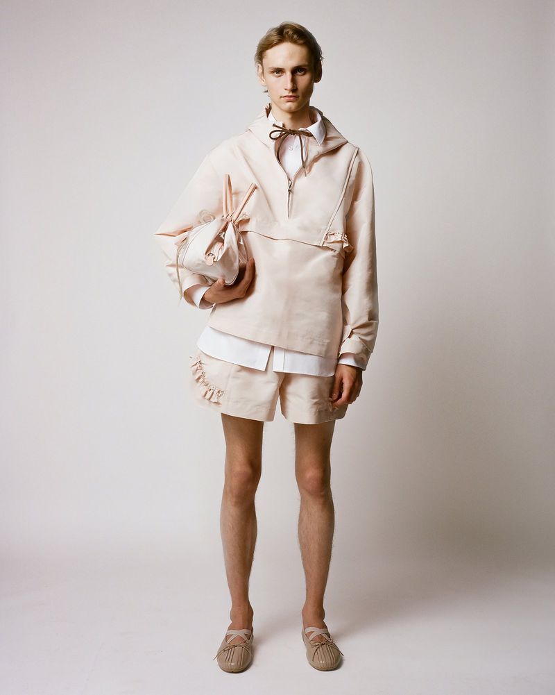 A slender male model poses in a studio wearing a tan linen anorak, shorts, and espadrilles, complemented by a matching linen bag.