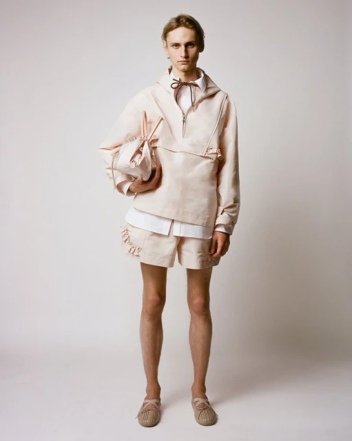 A slender male model poses in a studio wearing a tan linen anorak, shorts, and espadrilles, complemented by a matching linen bag.