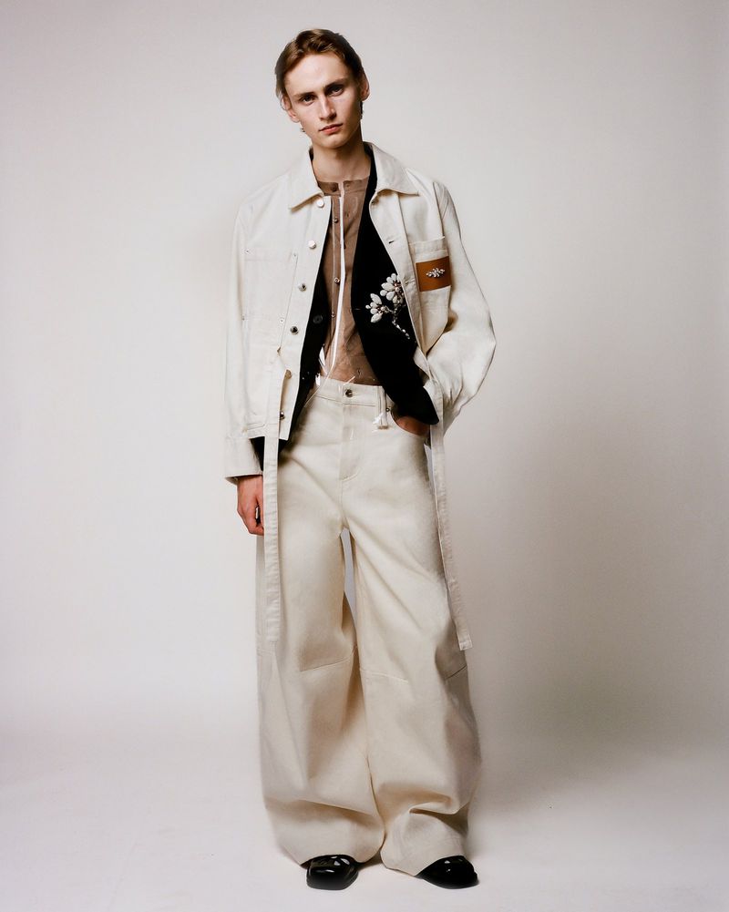 A young adult male poses in an off-white denim jacket and wide-leg trousers on a neutral backdrop in a fashion photography style.