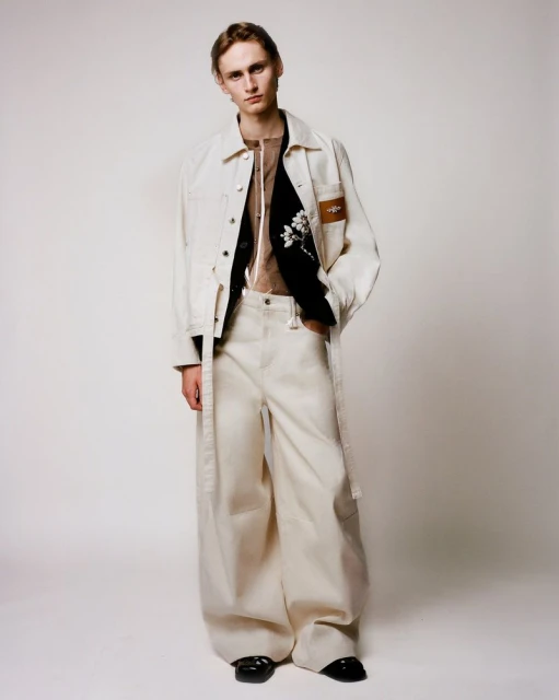 A young adult male poses in an off-white denim jacket and wide-leg trousers on a neutral backdrop in a fashion photography style.
