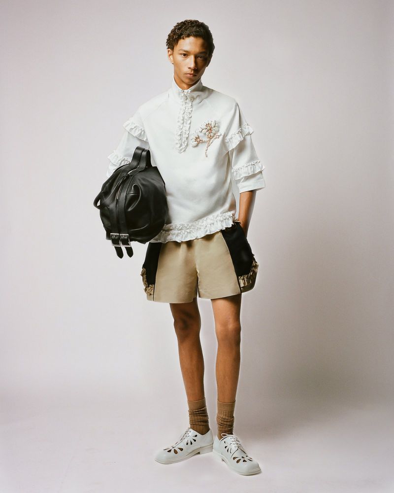 Full-shot fashion portrait of a slender young adult male, light skin, curly hair, posed holding a large black leather backpack on white.