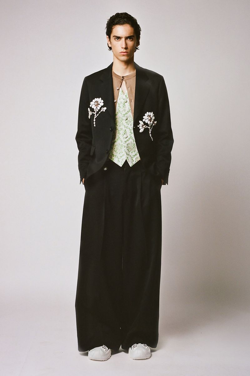 Full-length studio shot of a young male fashion model in a black jacket with floral embellishments, green patterned vest, and baggy trousers.