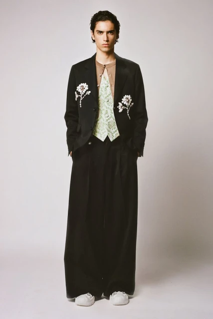 Full-length studio shot of a young male fashion model in a black jacket with floral embellishments, green patterned vest, and baggy trousers.