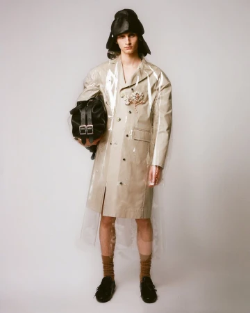 Fashionable young adult man in a transparent coat and black cap holds a black backpack against a solid light gray background.