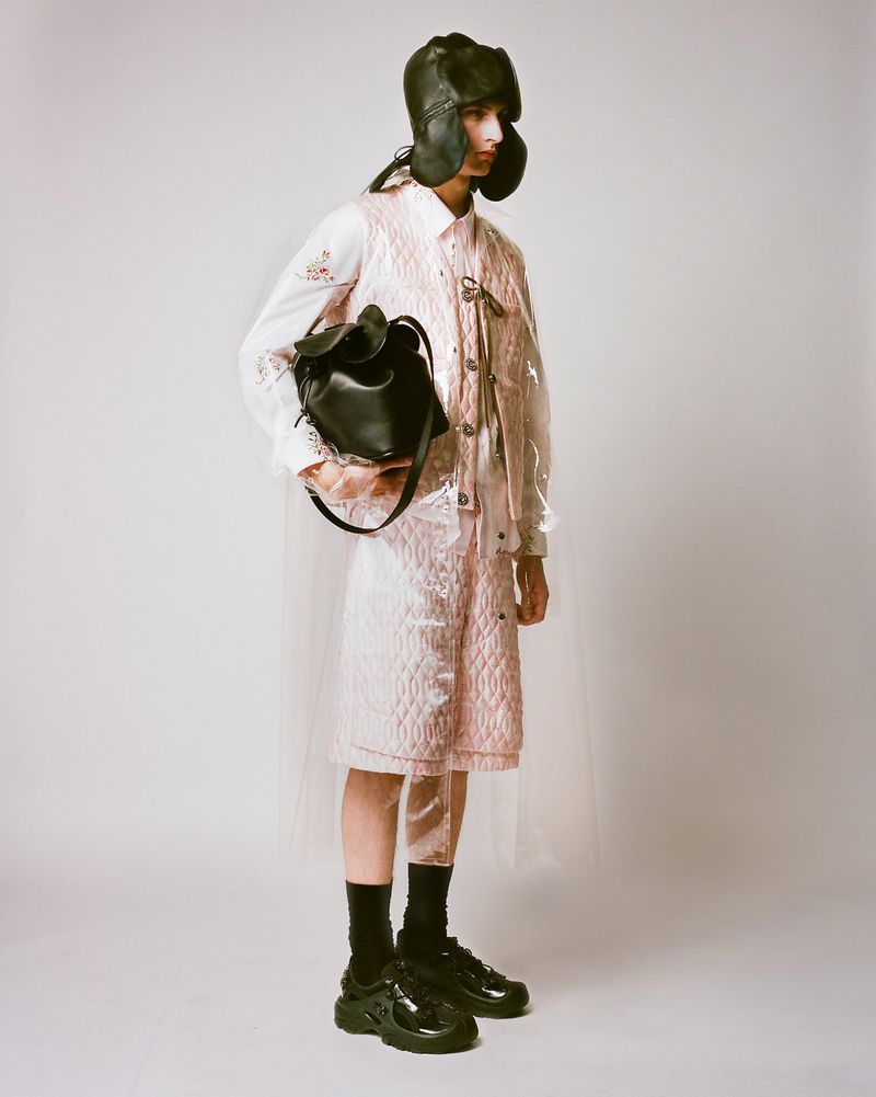 Fashionable person in studio wearing quilted outfit, balaclava hat, and black shoes, holding black bag against a neutral backdrop.