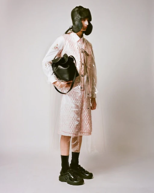 Fashionable person in studio wearing quilted outfit, balaclava hat, and black shoes, holding black bag against a neutral backdrop.