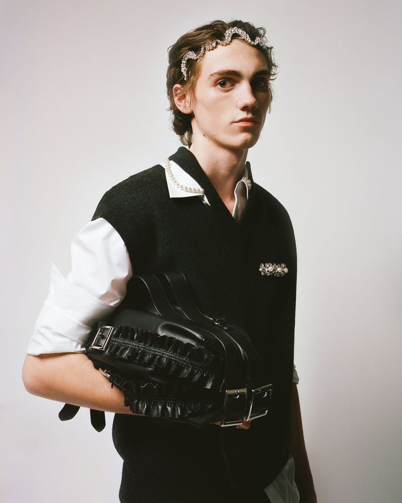 Adult male poses indoors wearing a black vest over a white shirt, jeweled headband, and holding a black ruffled handbag in studio lighting.