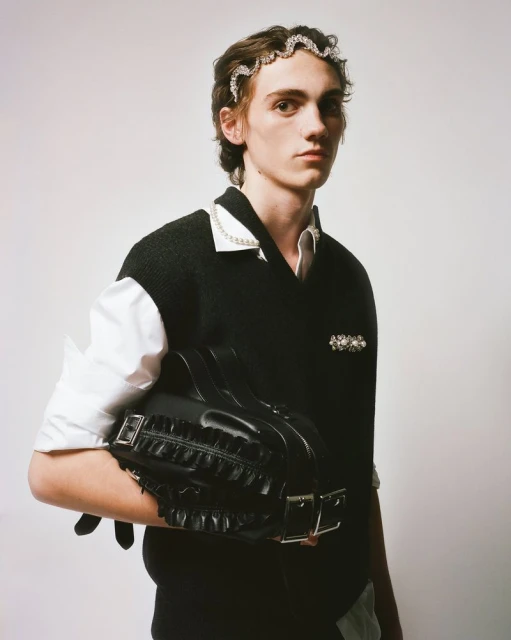 Adult male poses indoors wearing a black vest over a white shirt, jeweled headband, and holding a black ruffled handbag in studio lighting.