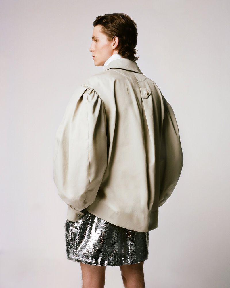 Back view of a male fashion model with light skin, wearing a tan jacket with large puffed sleeves and a silver sequined skirt in studio.