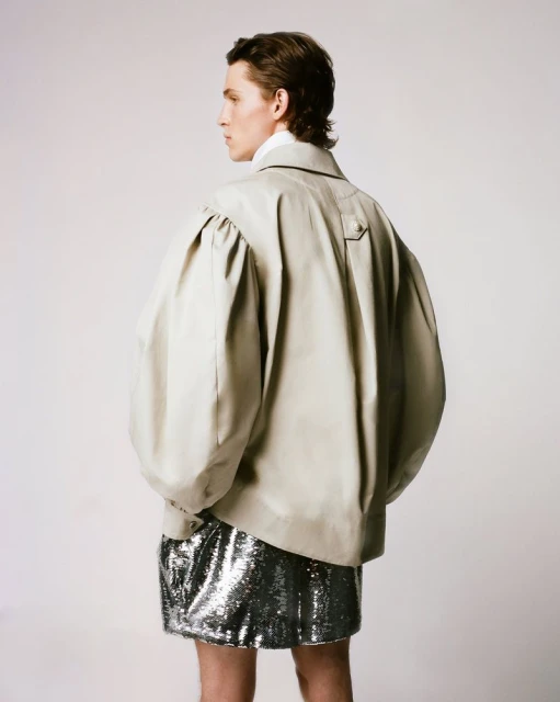 Back view of a male fashion model with light skin, wearing a tan jacket with large puffed sleeves and a silver sequined skirt in studio.