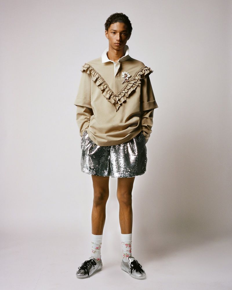 Full-length studio shot of a young adult with light-brown skin wearing a polo shirt, shorts, socks, and sneakers in front of a white background.