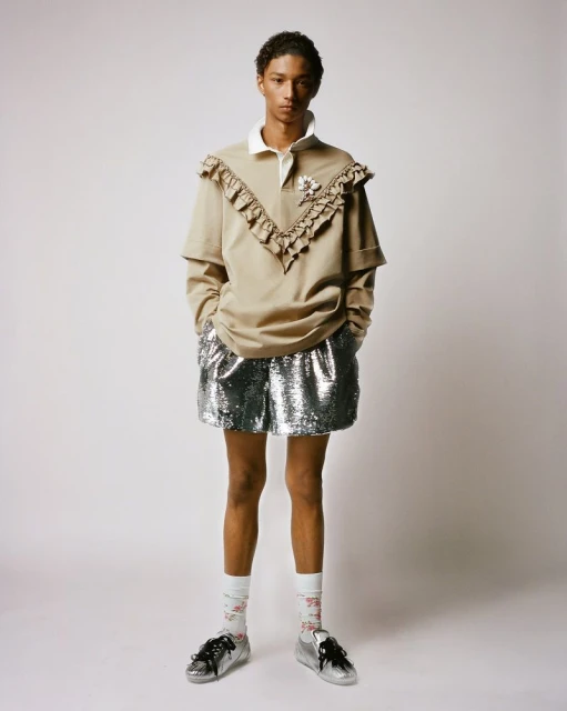 Full-length studio shot of a young adult with light-brown skin wearing a polo shirt, shorts, socks, and sneakers in front of a white background.