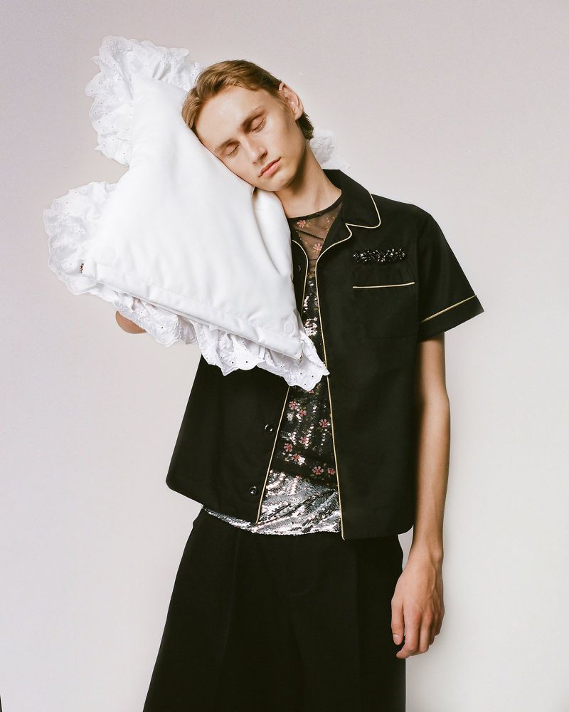 Young adult with pale skin and light brown hair poses indoors with closed eyes, wearing an unbuttoned shirt and holding a white pillow.