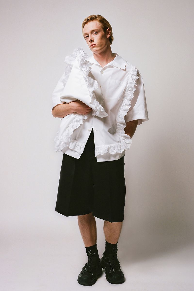 A fashionable red-haired male model poses in a white ruffled shirt and black shorts against a white backdrop in a studio setting.