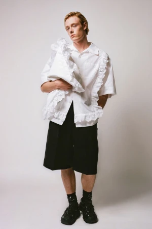 A fashionable red-haired male model poses in a white ruffled shirt and black shorts against a white backdrop in a studio setting.