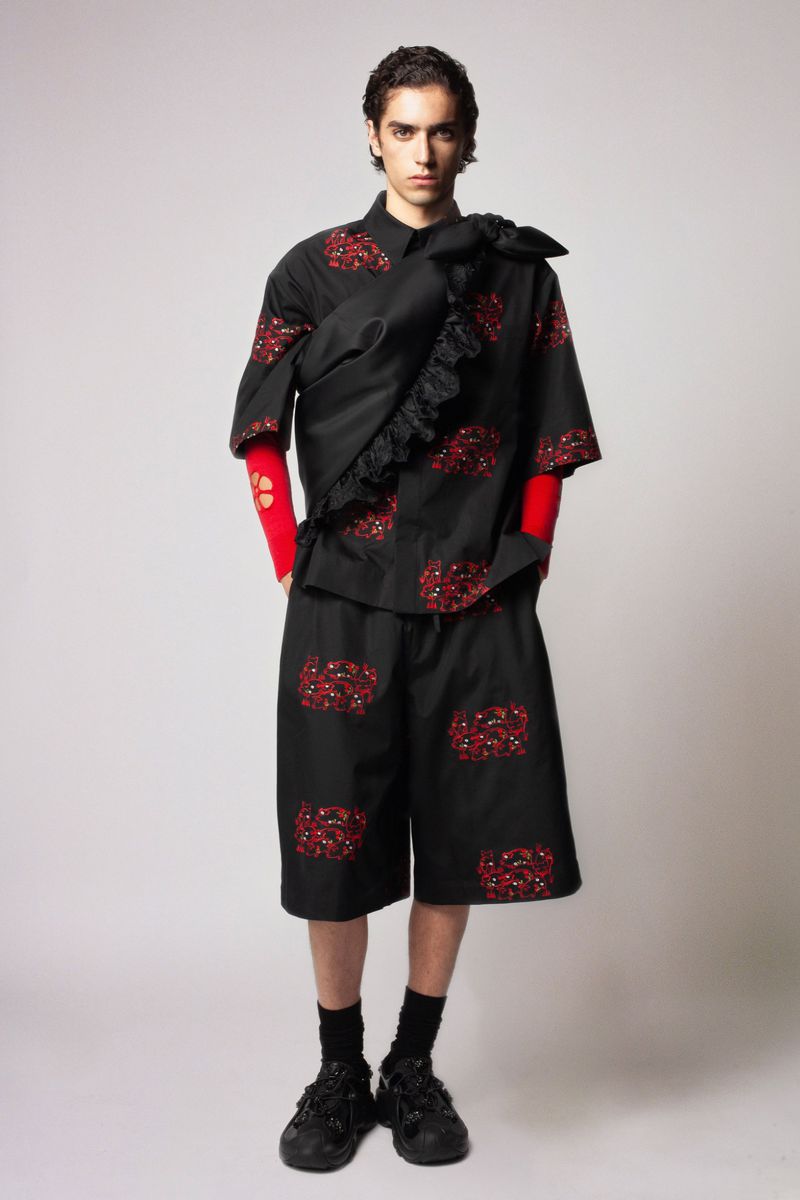 Fashion shot of a young adult man wearing a black patterned short-sleeve shirt and shorts, with black sneakers in a studio setting.