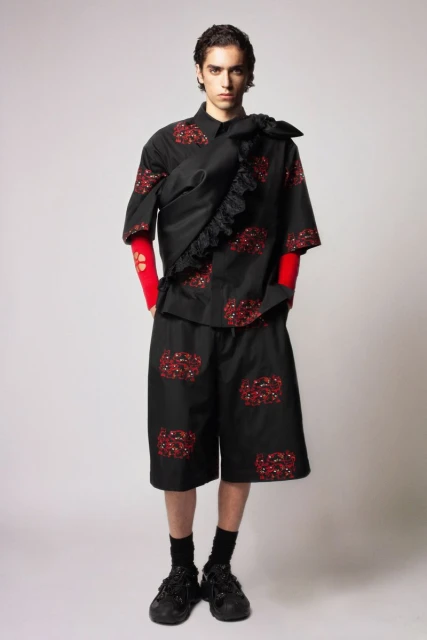 Fashion shot of a young adult man wearing a black patterned short-sleeve shirt and shorts, with black sneakers in a studio setting.