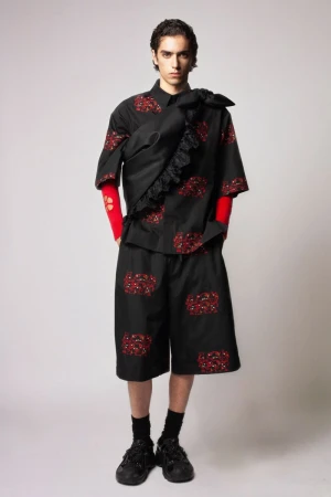 Fashion shot of a young adult man wearing a black patterned short-sleeve shirt and shorts, with black sneakers in a studio setting.