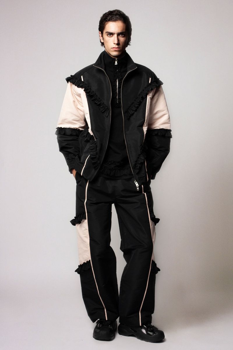 Full-length portrait of a young adult male in a black zip-up jacket and matching pants with pale pink accents against a plain background.