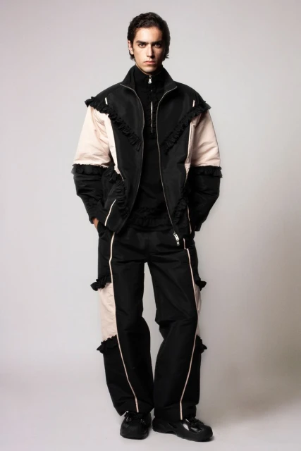 Full-length portrait of a young adult male in a black zip-up jacket and matching pants with pale pink accents against a plain background.