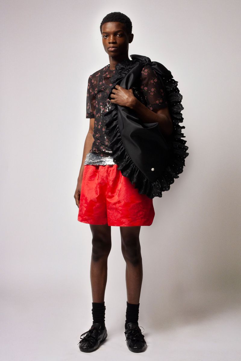 Full-length shot of a Black male model in a sheer floral shirt and red shorts, holding a ruffled black bag against a plain background.
