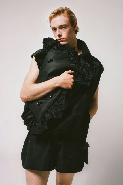 Young adult posing in black sartorial vest and shorts against a white backdrop, indoor studio lighting, fashion forward creative direction.