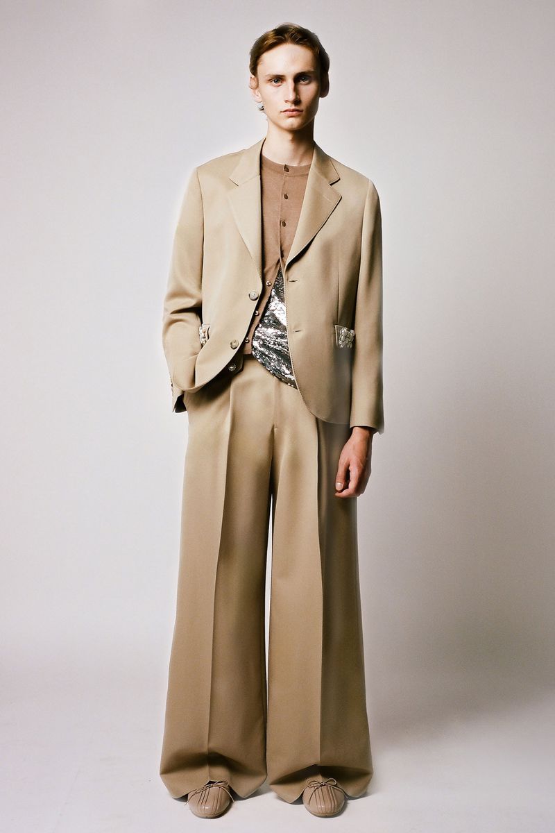 A person stands in a photo studio wearing a tan blazer and wide-legged trousers. The subject is wearing a brown shirt and beige shoes.