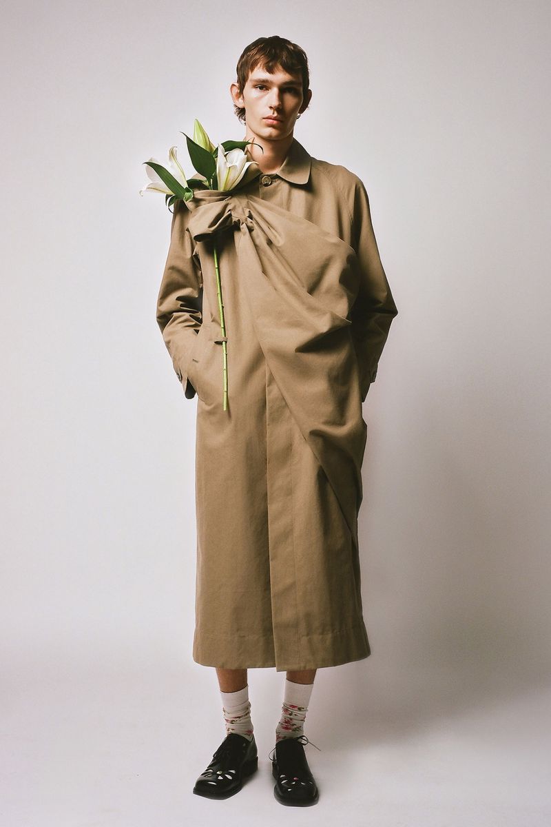 Adult male fashion model wearing a tan trench coat with gathered fabric and carrying white lilies poses indoors against a white backdrop.