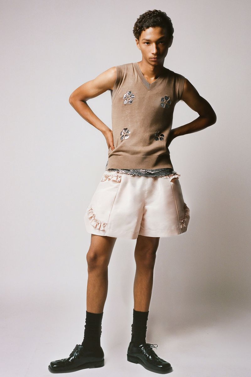Fashion shot of a young male adult wearing a tan tank top and light pink ruffled shorts with black socks and loafers on white backdrop.