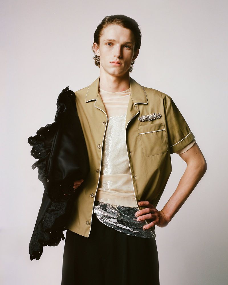 Adult male fashion model wearing a tan short-sleeve shirt with black skirt and jacket, posing in a studio with an off-white background.