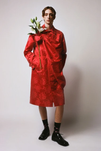 Man poses in a red floral coat holding white lilies in a studio setting, wearing shiny black shoes, with soft lighting.