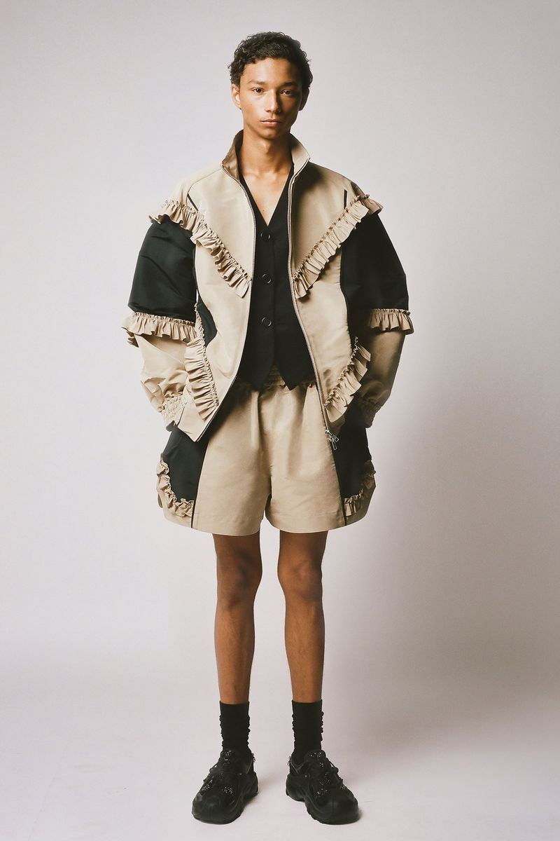 Full-length portrait of a slender person with dark hair wearing a tan jacket and shorts set, standing in front of a light background.