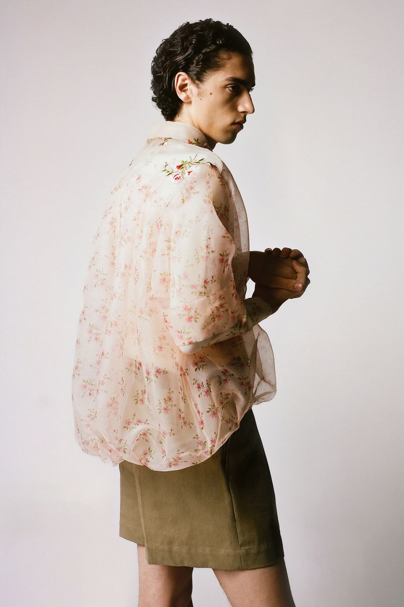 Side shot of a slender man with curly hair wearing a floral translucent jacket and khaki shorts in a brightly lit studio.