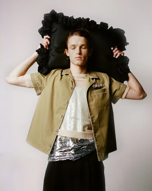 Adult male poses with a black, frilled pillow, wearing a tan button-up shirt, sequined top and belt, and dark pants against a light background.