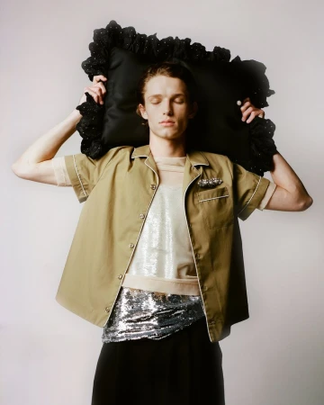 Adult male poses with a black, frilled pillow, wearing a tan button-up shirt, sequined top and belt, and dark pants against a light background.