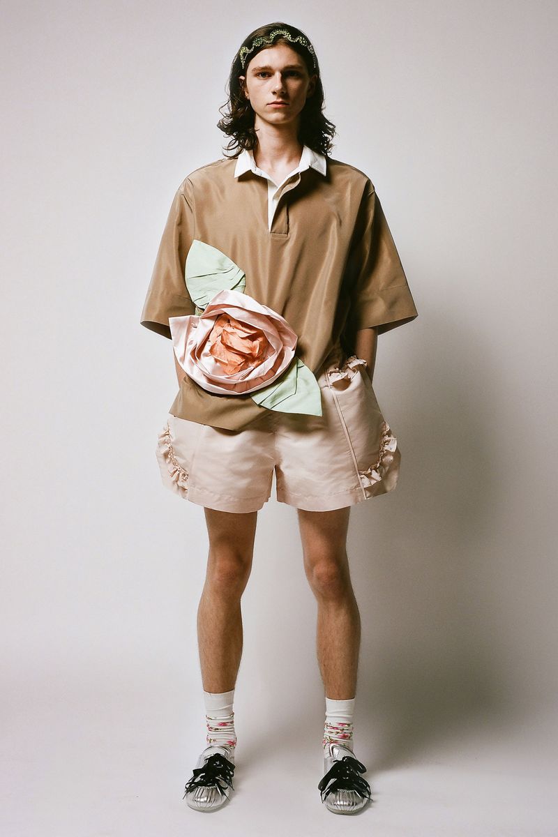 Full-length portrait of a young adult with long hair wearing a tan polo shirt with an oversized flower, pale pink shorts, and embellished sandals.