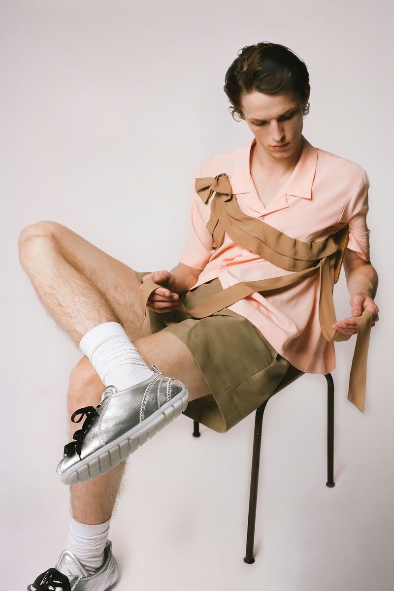 A young fair-skinned man is sitting on a stool with silver sneakers, a peach-colored shirt, and green shorts in a studio setting.