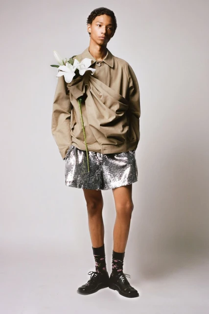 Full-length portrait of a male model in a tan jacket and silver shorts, accessorized with flowers, against a neutral backdrop.