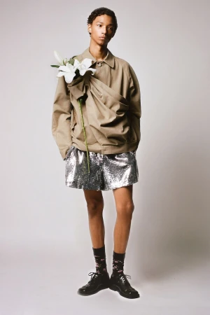 Full-length portrait of a male model in a tan jacket and silver shorts, accessorized with flowers, against a neutral backdrop.