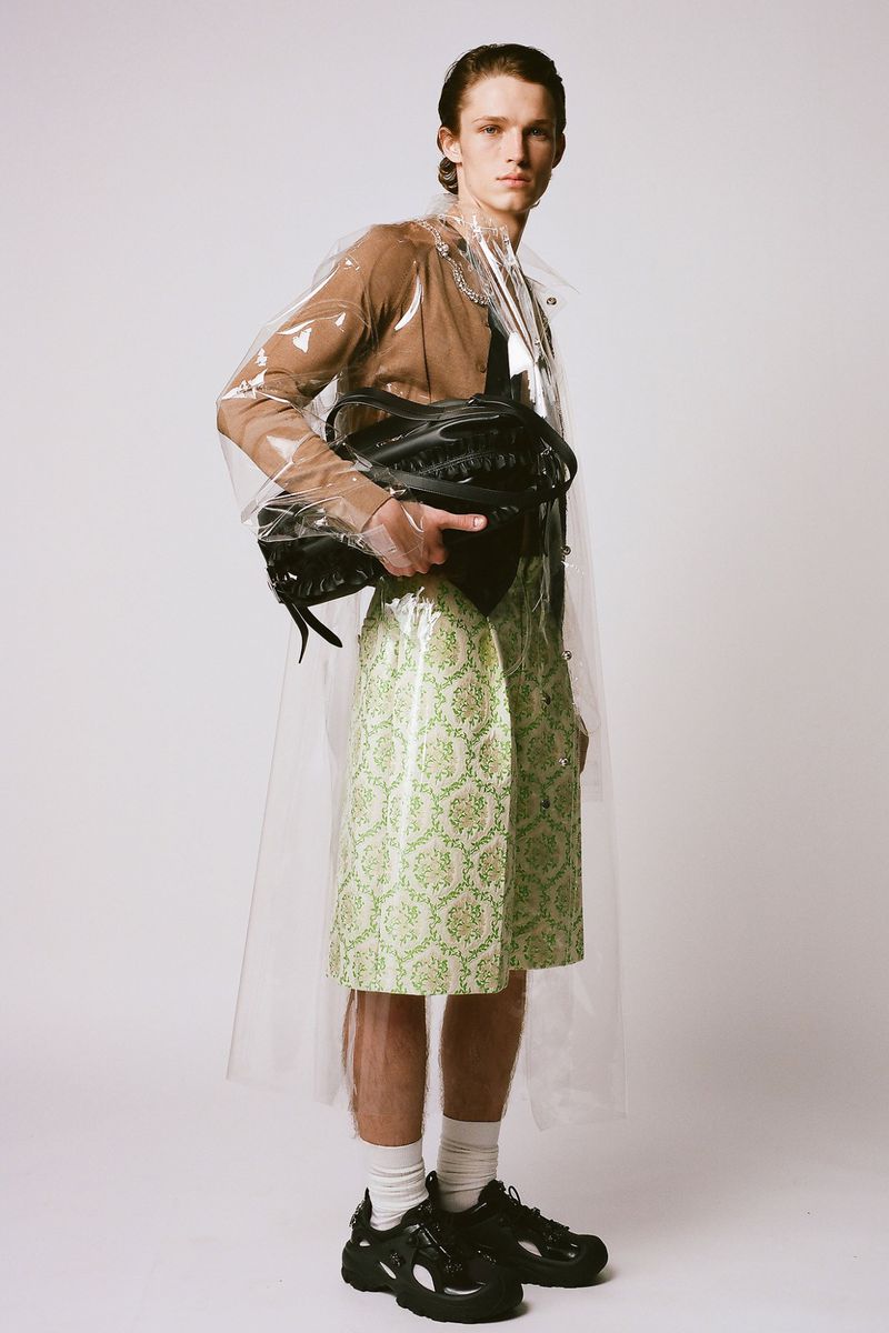 A slender man poses in a translucent raincoat, brown top, knee-length green patterned skirt, white socks, and black sneakers on a white background.