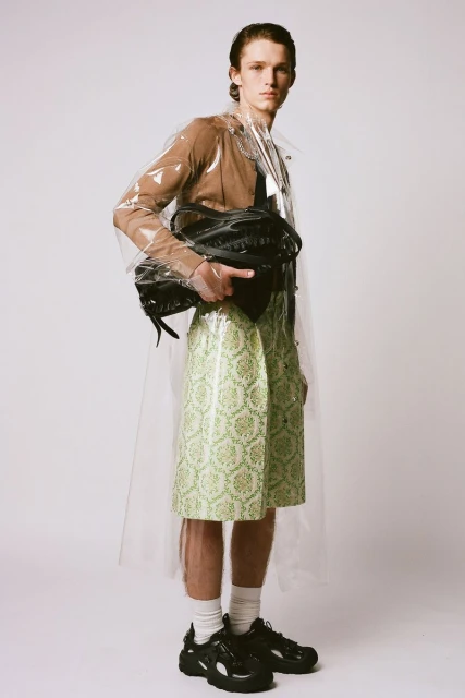 A slender man poses in a translucent raincoat, brown top, knee-length green patterned skirt, white socks, and black sneakers on a white background.