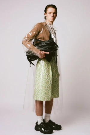 A slender man poses in a translucent raincoat, brown top, knee-length green patterned skirt, white socks, and black sneakers on a white background.