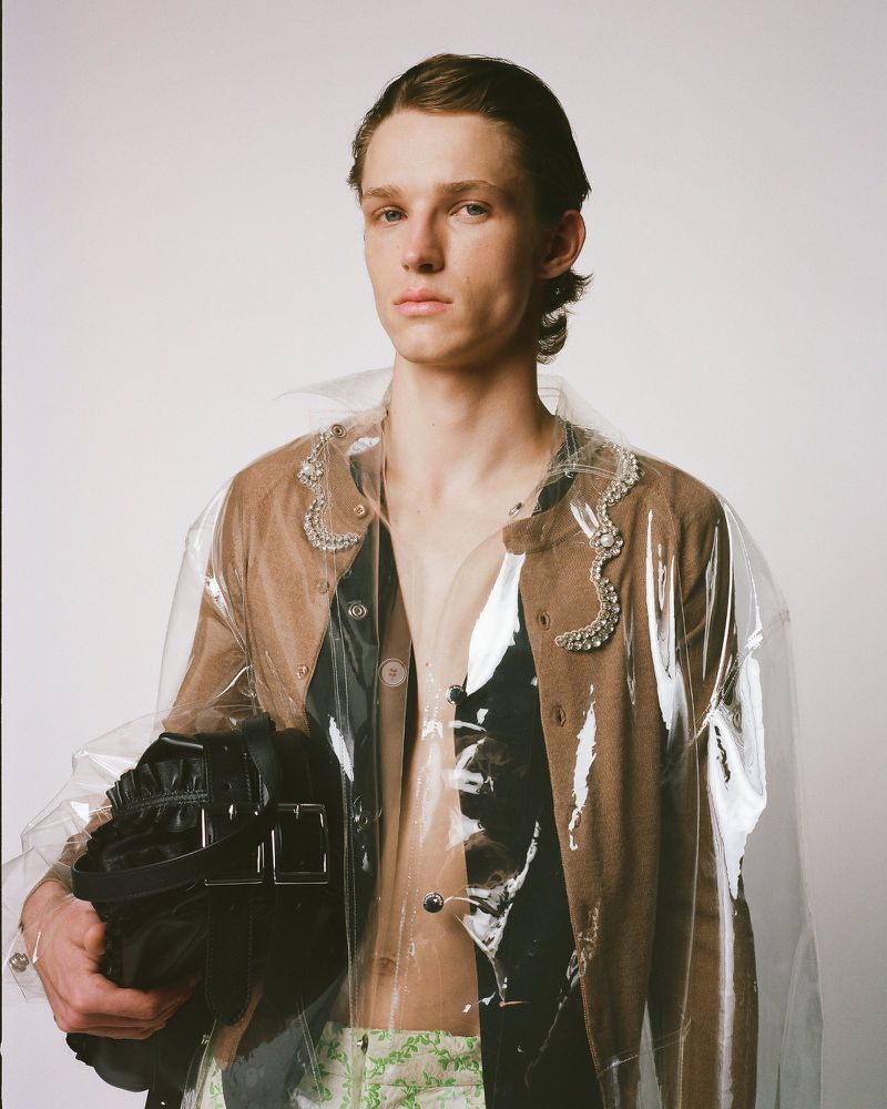 Man in brown cardigan wearing a clear plastic coat holds a black bag in a studio setting against a plain white background.