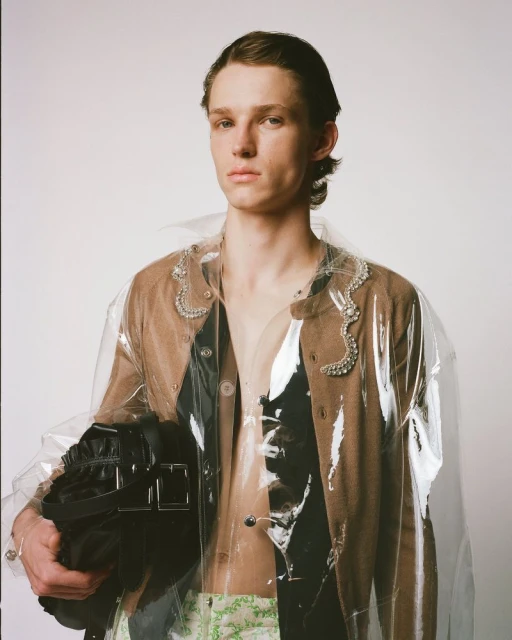 Man in brown cardigan wearing a clear plastic coat holds a black bag in a studio setting against a plain white background.